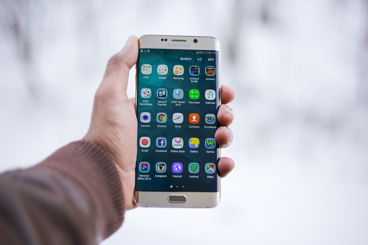 A hand holding a Samsung smartphone displaying a variety of apps on the screen, with a blurred white background.